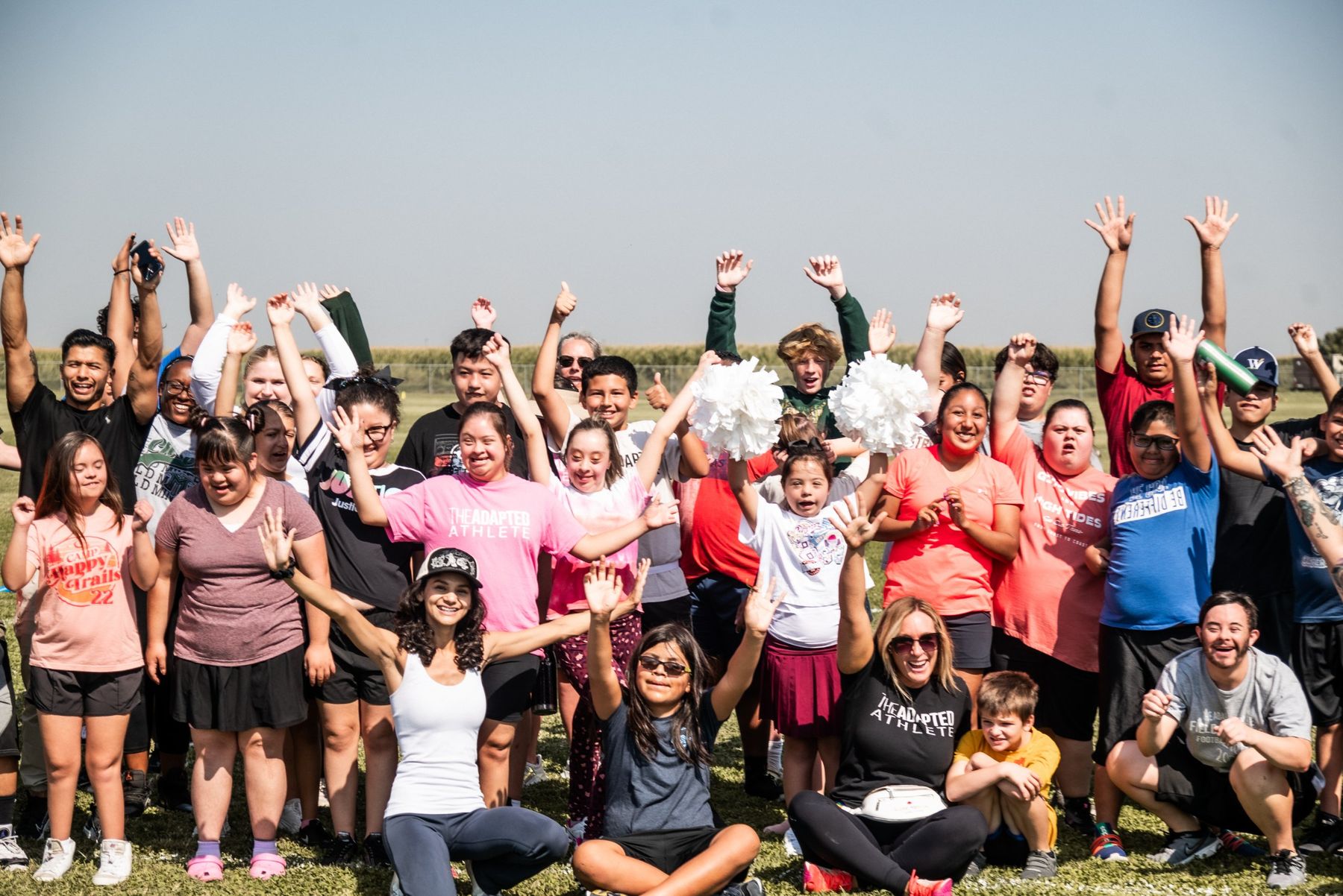 A big group of athletes, coaches, and families cheering with arms raised on a sunny field